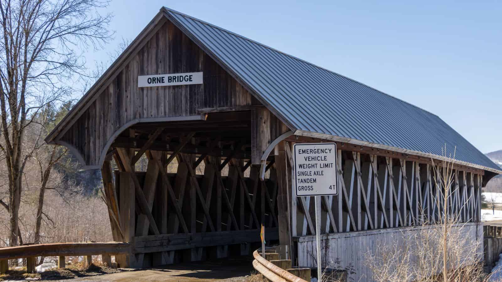 A wooden covered bridge labeled "Orne Bridge" with a metal roof, next to a sign stating emergency vehicle weight limits; trees and hills are visible in the background.