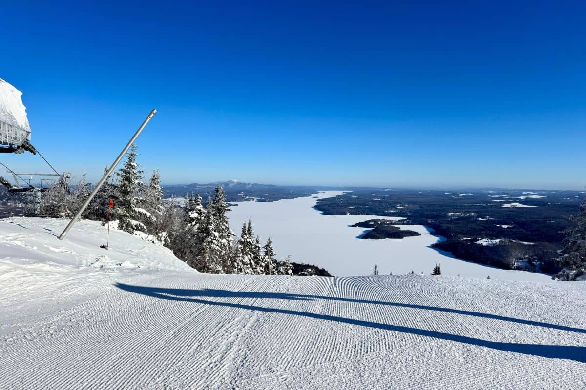 Snow-covered ski slope overlooking a frozen lake and forested landscape under a clear blue sky.