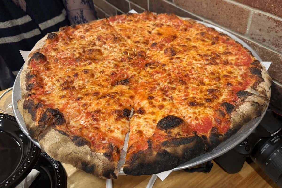 A large, thin-crust cheese pizza with a charred, crispy edge is served on a metal tray in a restaurant setting.
