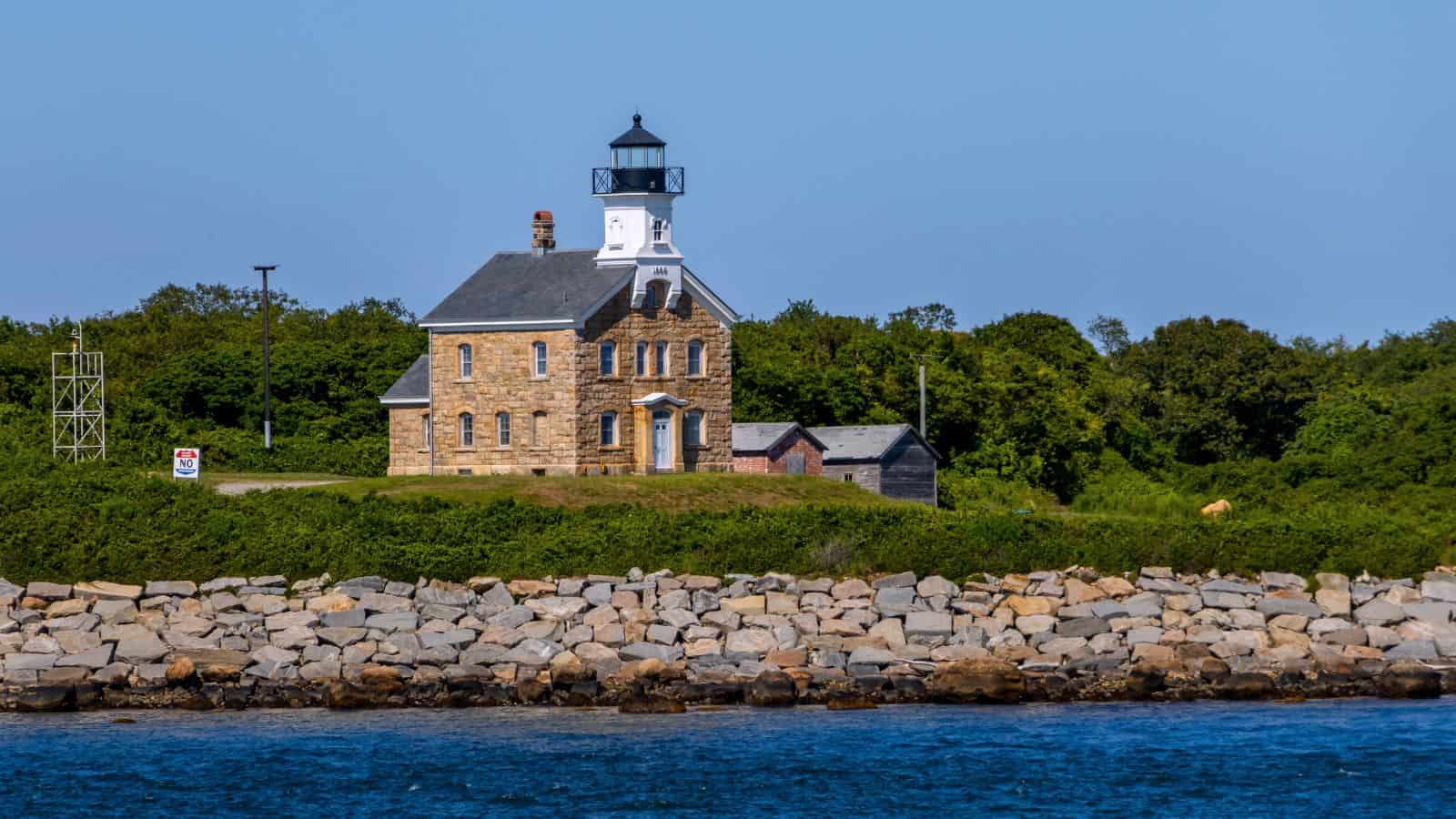 A stone lighthouse with an attached house stands near the shore, surrounded by greenery and rocks, under a clear blue sky.