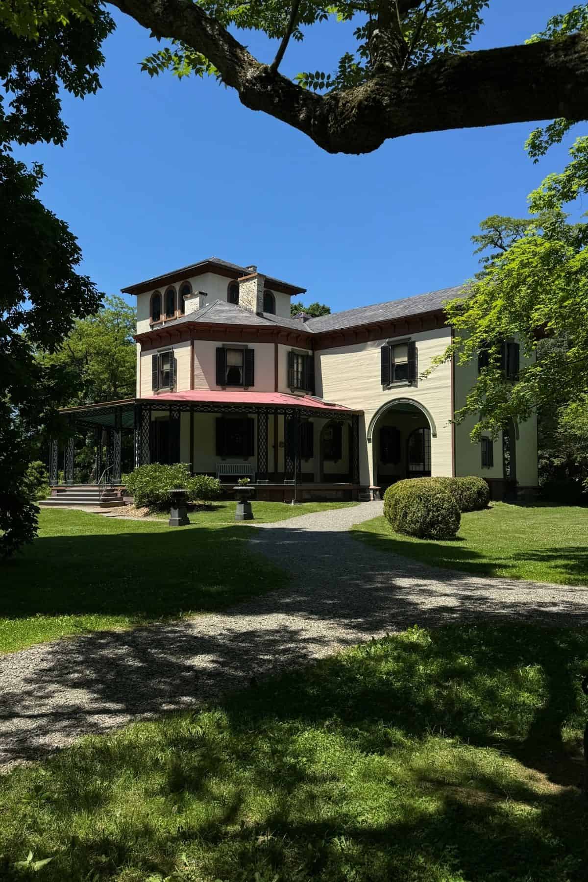 A large, cream-colored Victorian house with a red roof sits on a grassy lawn, surrounded by trees and a gravel path under a clear blue sky.