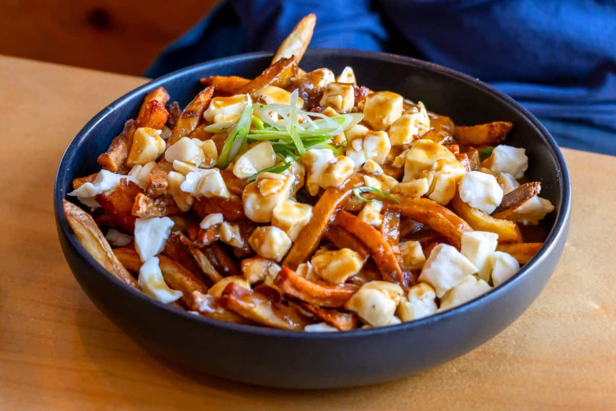 A black bowl filled with poutine, featuring French fries topped with cheese curds, brown gravy, and garnished with sliced green onions, on a wooden table.