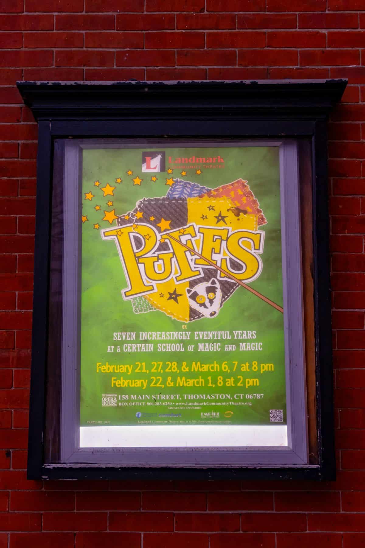 A theater poster for "Puffs" is displayed in a black frame on a red brick wall, advertising show dates in February and March at Landmark Community Theatre in Thomaston, CT.