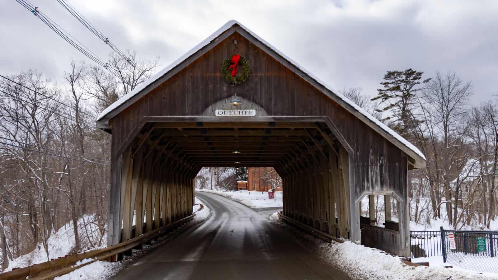 A wooden covered bridge with a wreath hangs above the entrance; snow covers the ground and trees, and a road runs through the bridge.