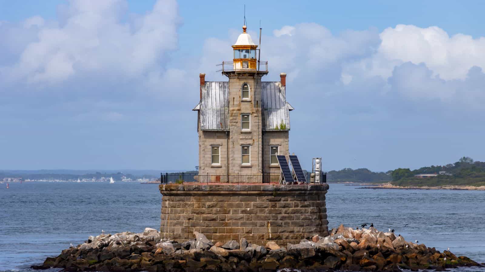A stone lighthouse with a square base and metal roof sits on a rocky platform surrounded by water, with solar panels and distant land visible in the background.