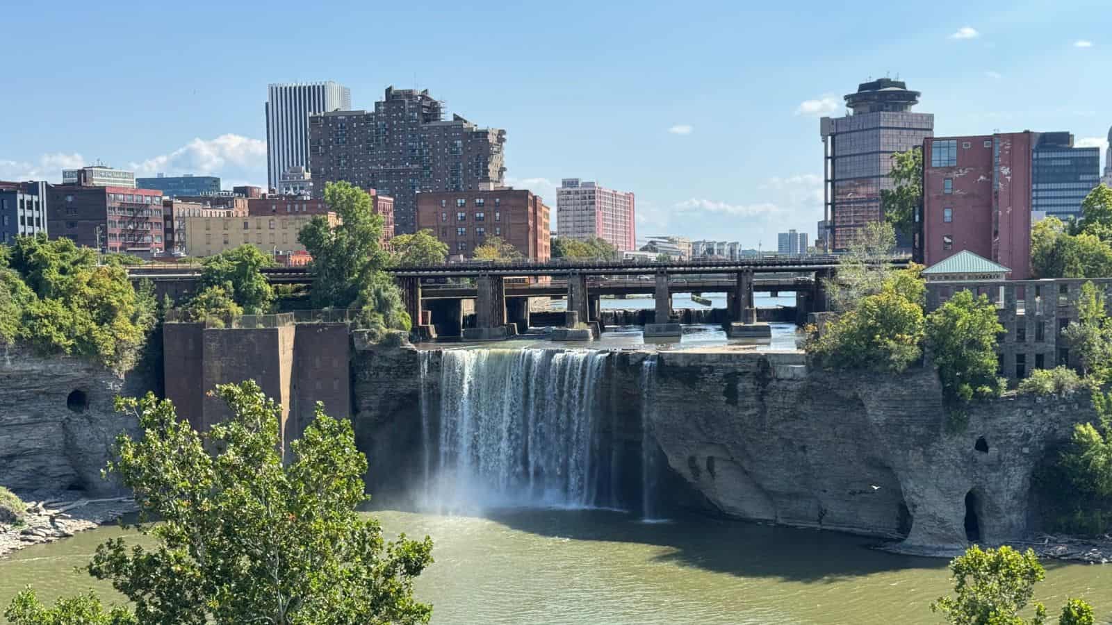 A waterfall flows over a rocky ledge into a river, with city buildings and a bridge in the background under a clear sky.