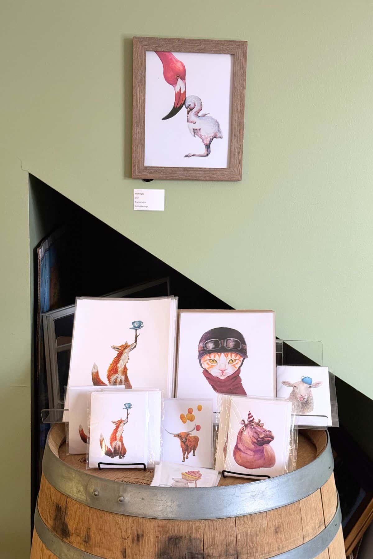 A framed illustration of two birds hangs above a display of animal-themed art prints arranged on a wooden barrel against a pale green wall.