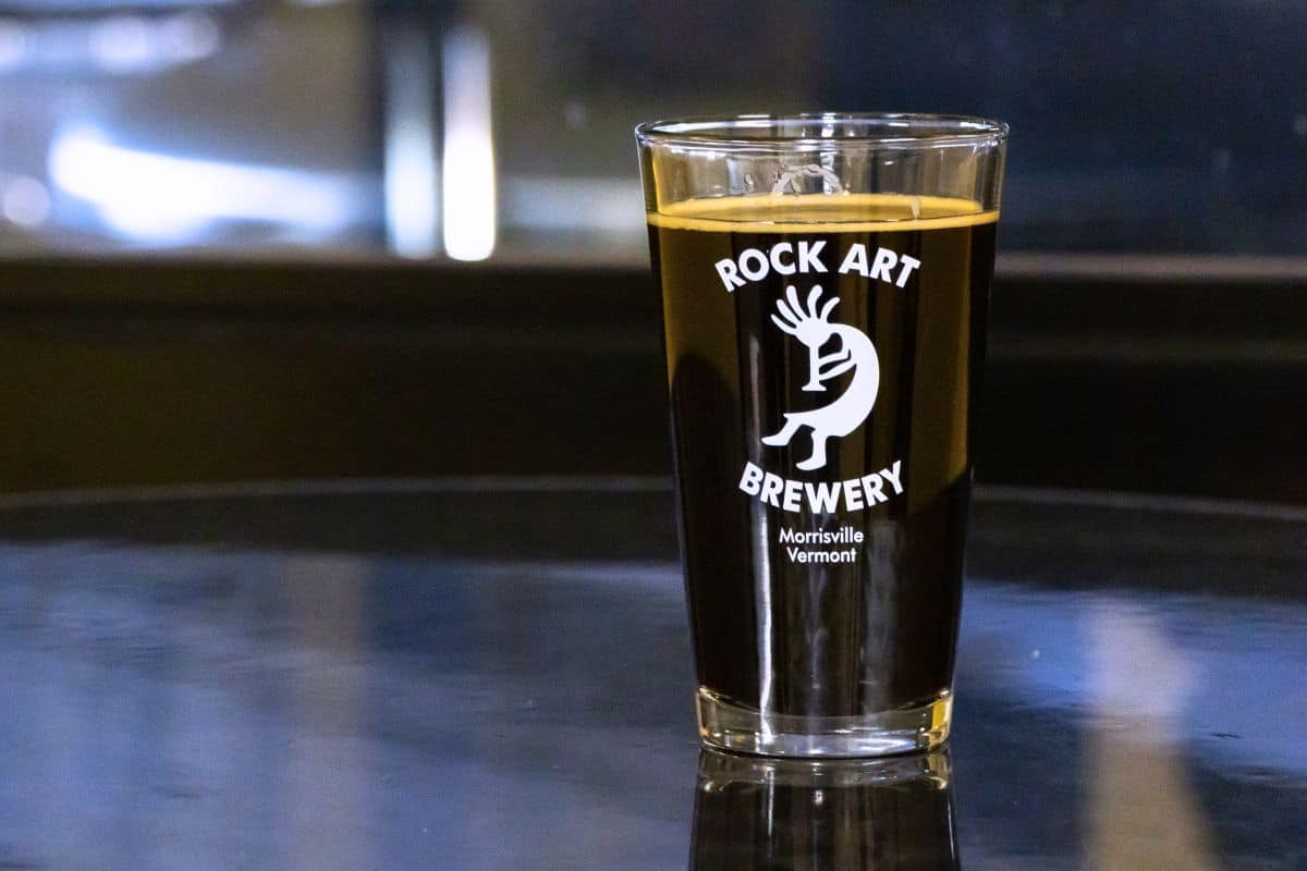 A pint glass filled with dark beer sits on a reflective surface. The glass is branded with "Rock Art Brewery, Morrisville Vermont" and a stylized figure logo.