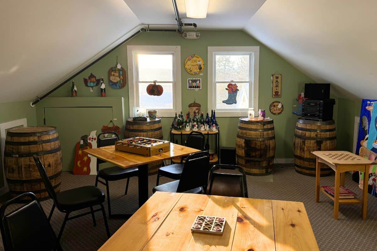 Room with slanted ceiling, wooden tables, chairs, barrels, board games, and various decorations on light green walls near two windows.