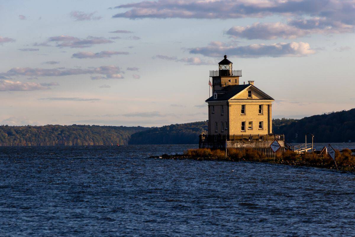 A lighthouse with a square base stands on the edge of a calm lake, with distant hills and a partly cloudy sky in the background.