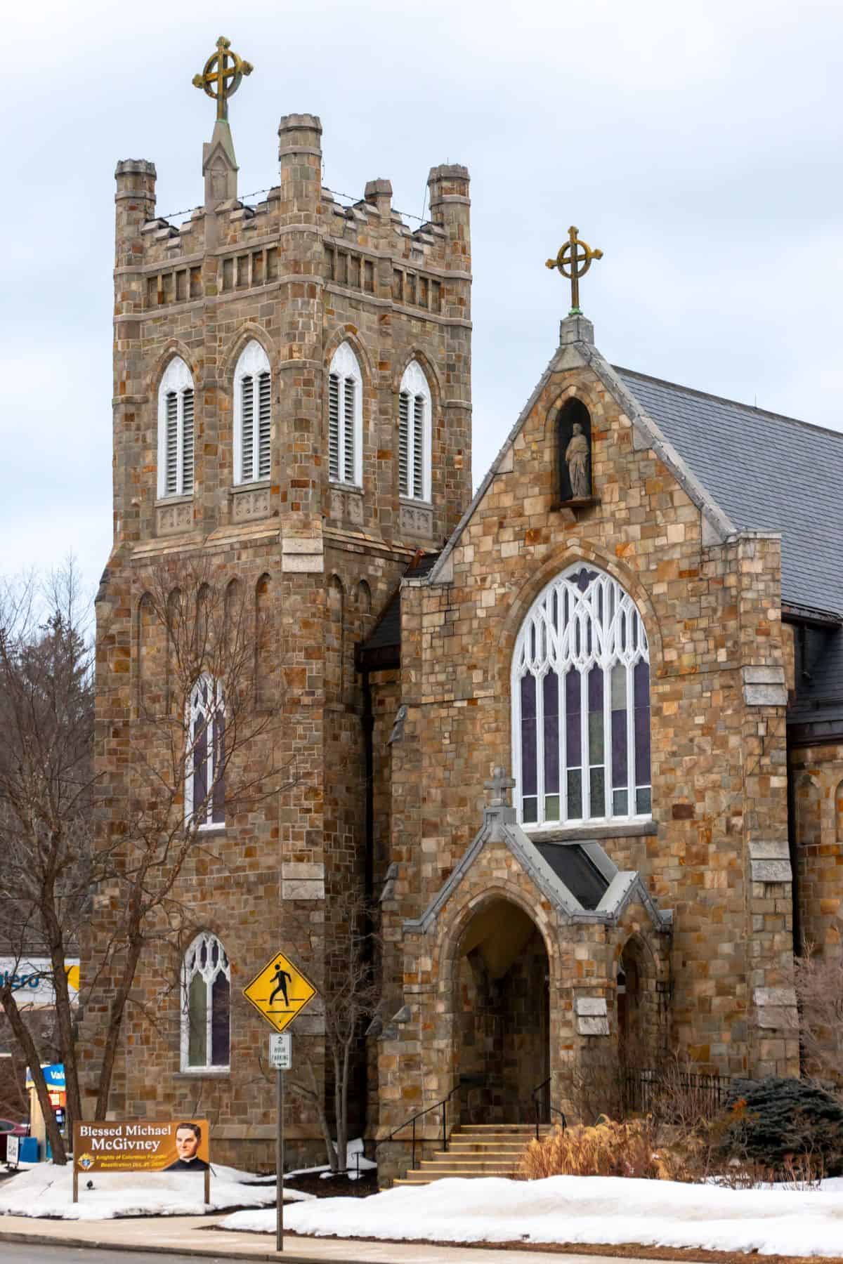Stone Gothic-style church with arched windows, a tall tower, and cross finials, set against a cloudy sky with snow on the ground and a pedestrian crossing sign in front.