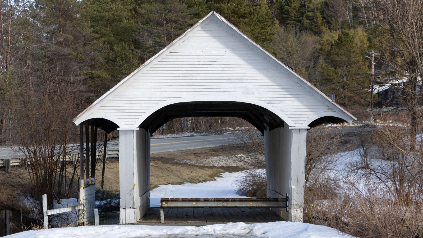 A white covered wooden bridge stands over a small creek with some snow on the ground and bare trees nearby; a road runs parallel in the background.