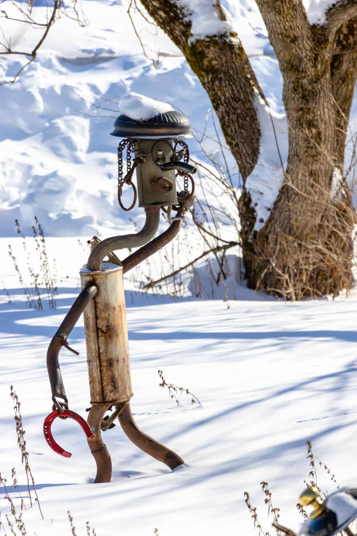 A sculpture made of metal scrap parts, shaped like a person, stands in snow near leafless trees, holding a red horseshoe.