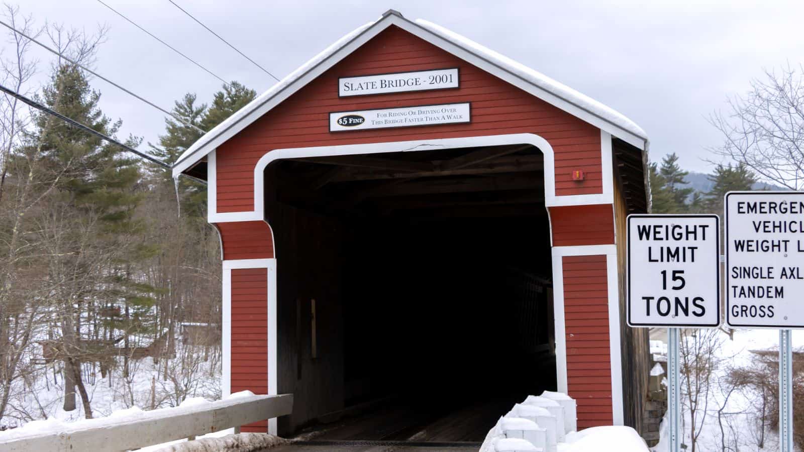 A red covered bridge with a sign reading "Slate Bridge 2001" and nearby road signs indicating a 15-ton weight limit in a snowy landscape.