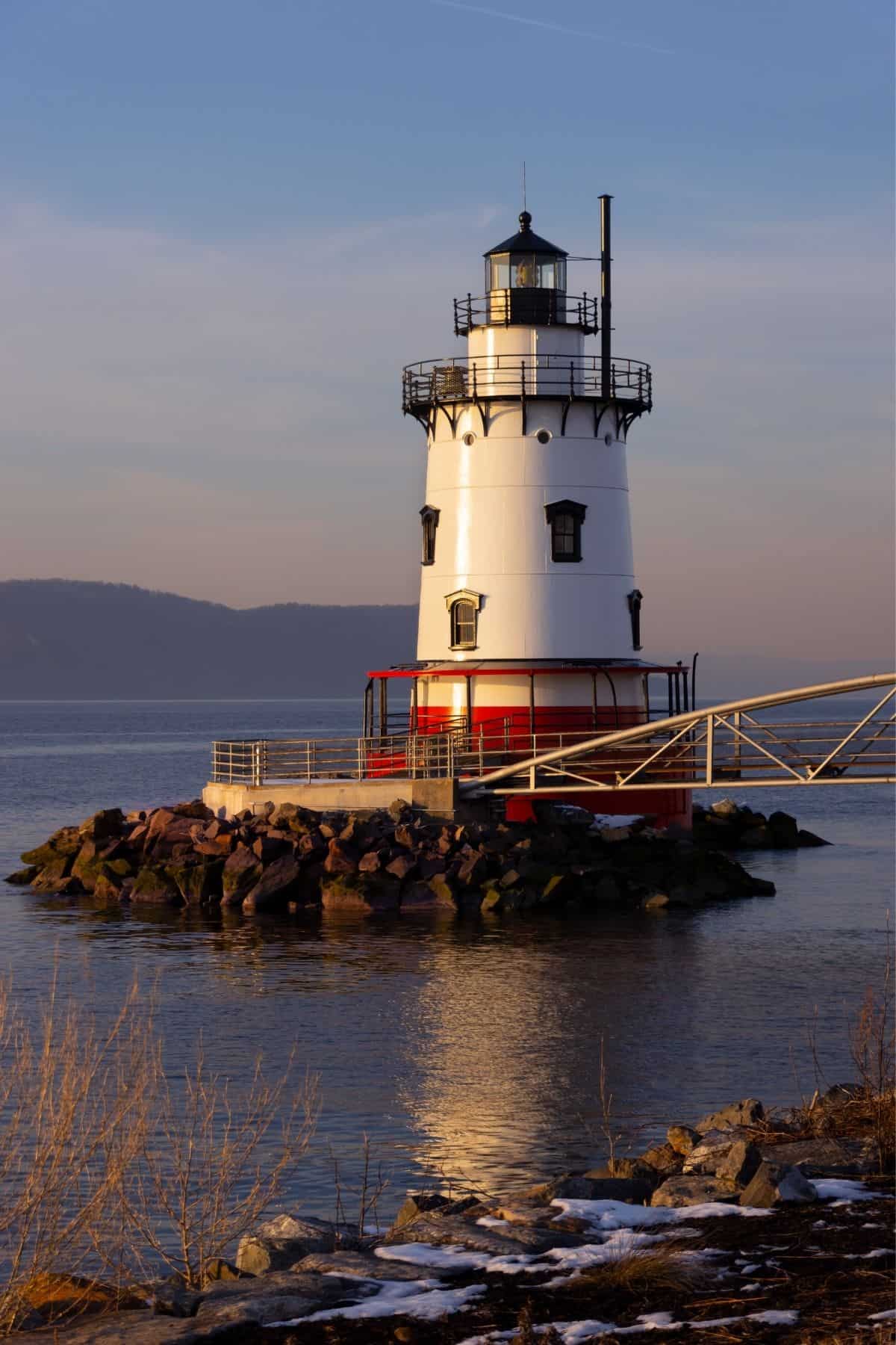 A white and red lighthouse stands on a rocky shore surrounded by water at sunset, with hills in the background and a metal walkway leading to the structure.