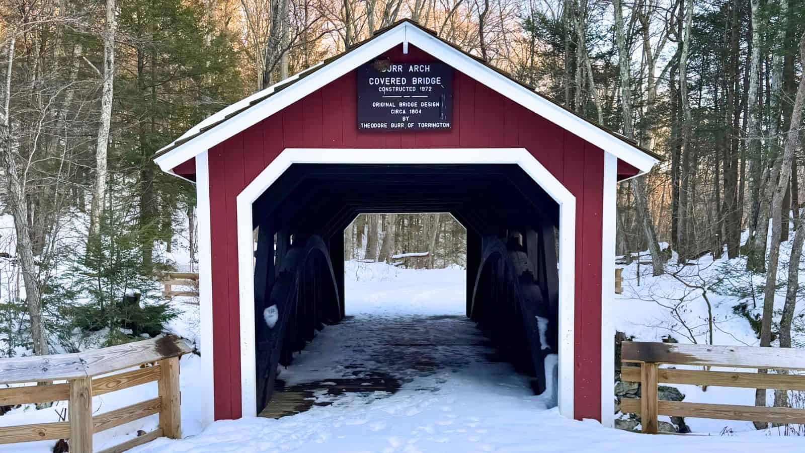A red covered bridge with a wooden sign stands over a snowy path, surrounded by trees in a winter landscape.