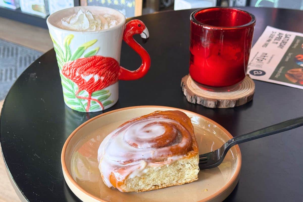 A frosted cinnamon roll on a plate with a fork next to a mug with a flamingo handle and a red glass candle holder on a round black table.
