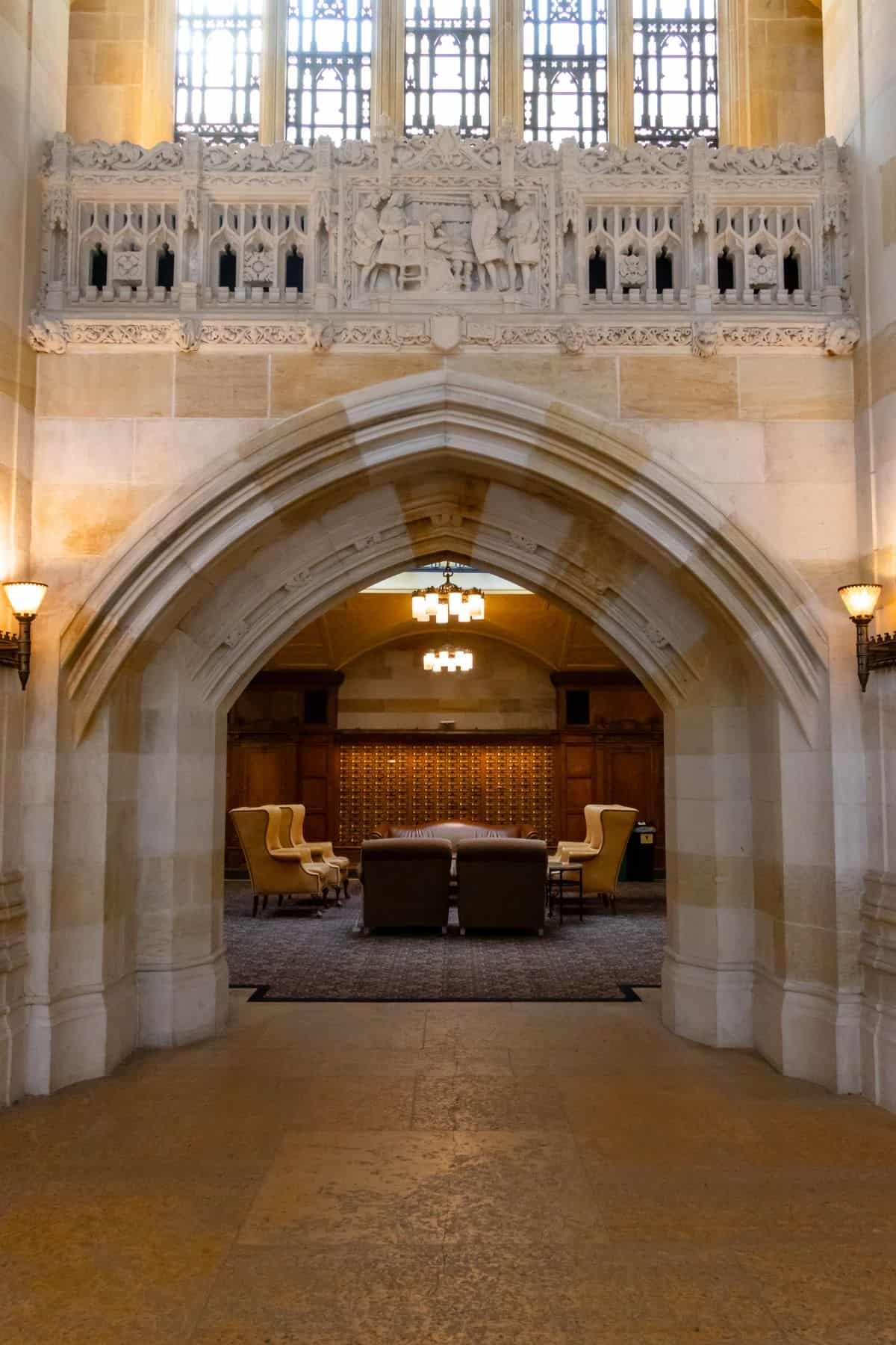 A stone archway leads into a room with armchairs, a sofa, wooden paneling, and a patterned rug, illuminated by chandeliers and wall sconces.