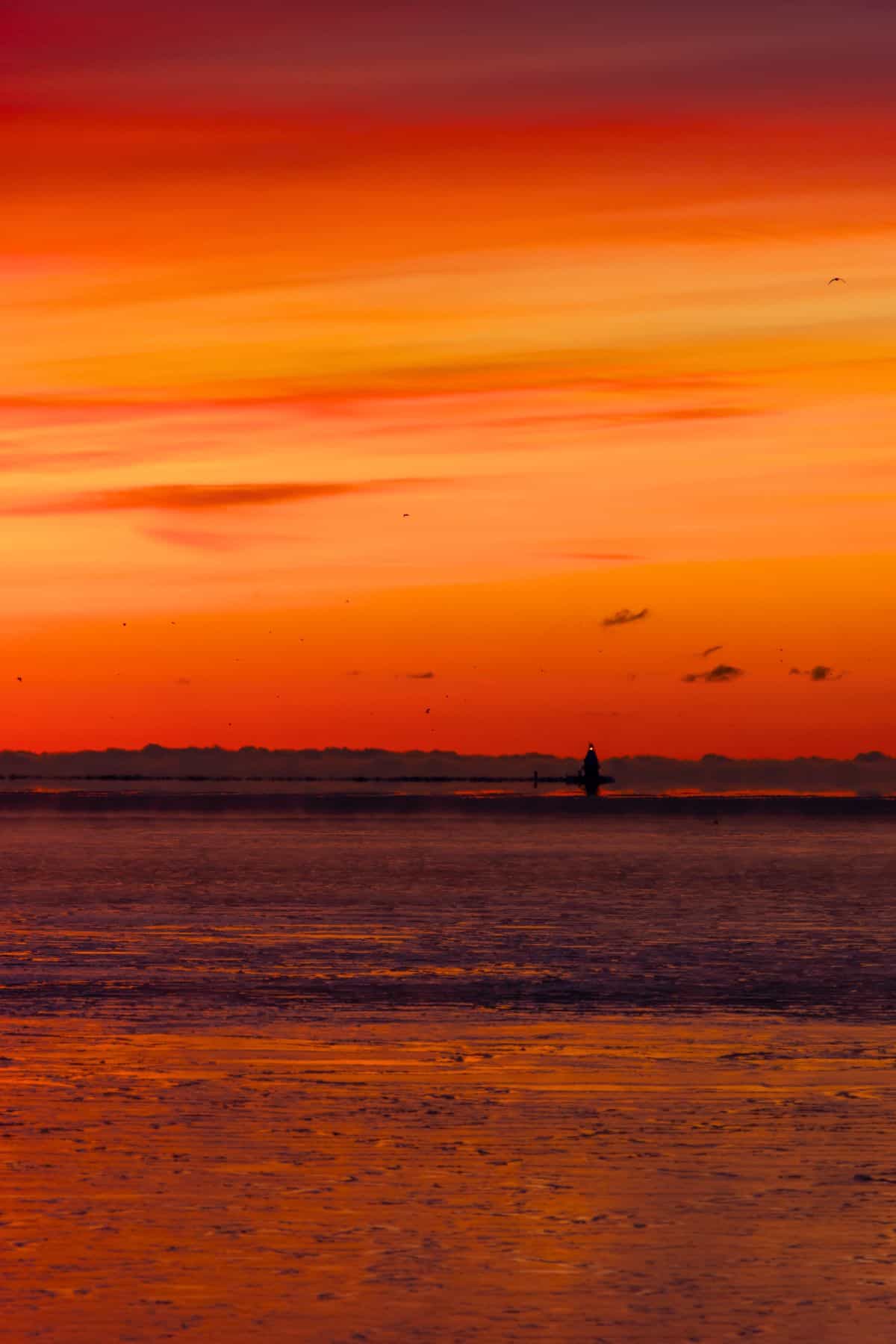 A vibrant sunset sky with orange and red hues reflects on the calm water, with a small silhouette of a buoy or lighthouse in the distance.