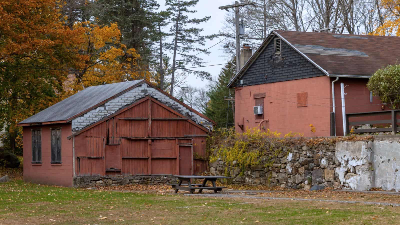 Two rustic red buildings with weathered roofs are situated next to a stone wall, surrounded by autumn trees and a picnic table on a grassy area.