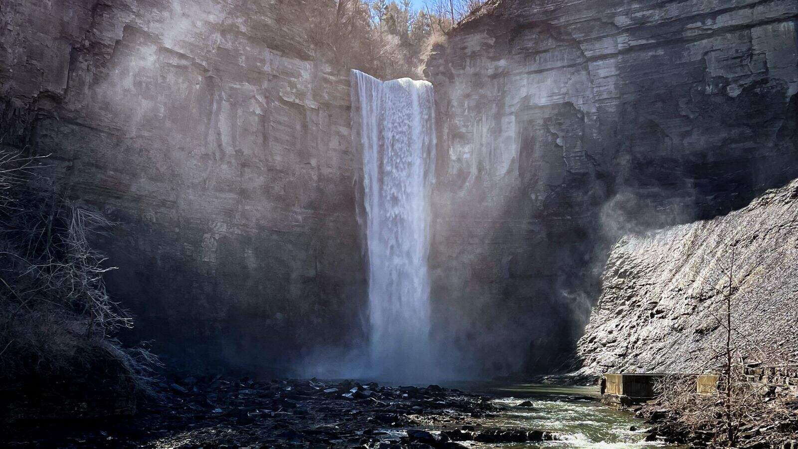 A waterfall cascades down a rocky cliff into a pool below, surrounded by steep rock walls and sparse vegetation under daylight.