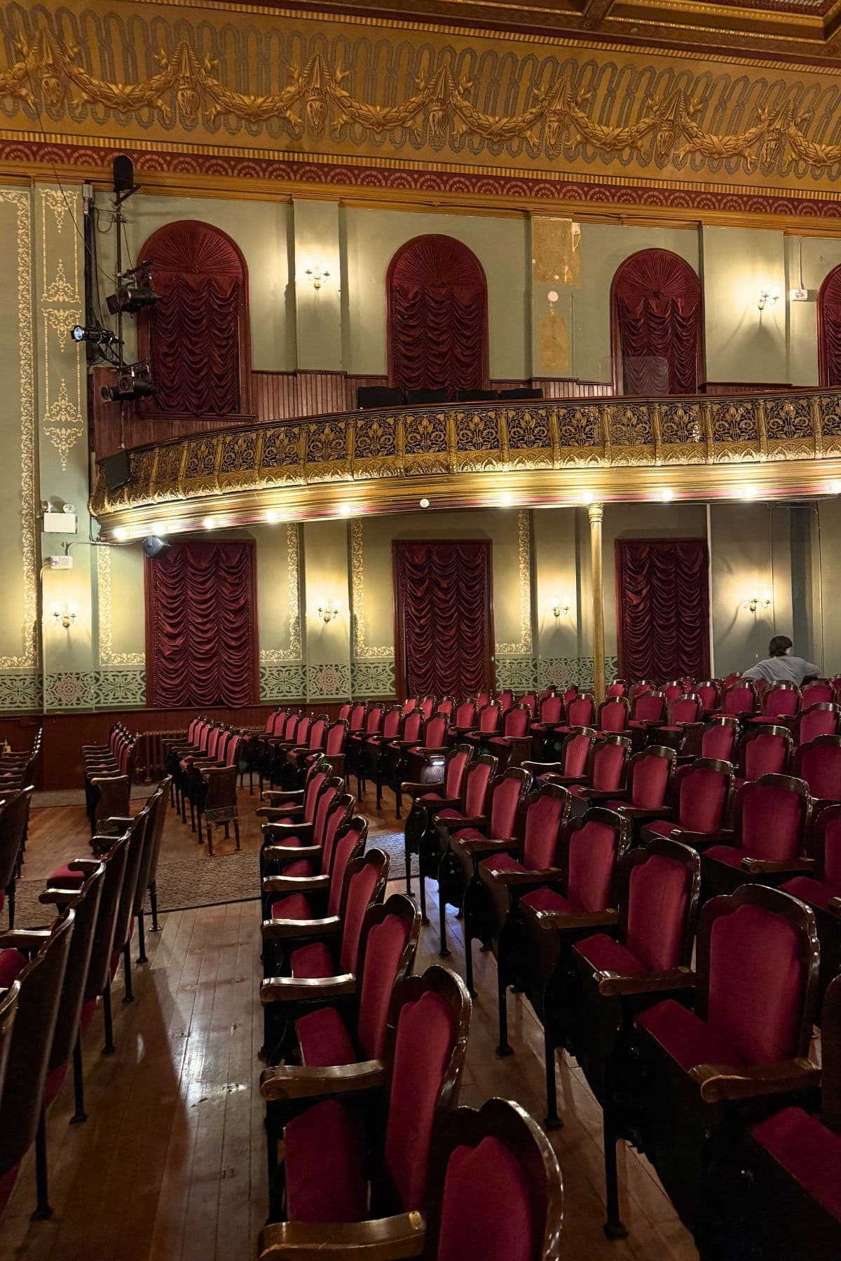 Rows of empty red velvet seats in an ornate theater with gold detailing, chandeliers, and a decorative balcony.