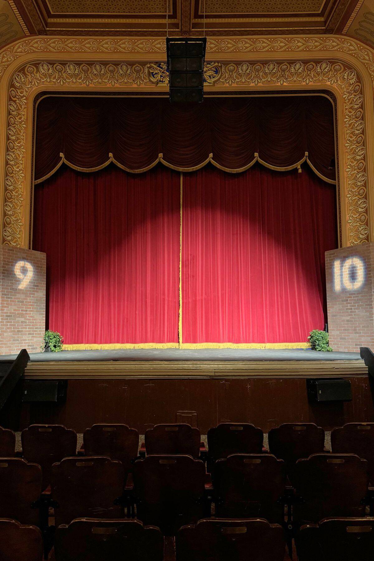 A theater stage with closed red curtains, gold trim, and brick columns on each side displaying the numbers 9 and 10, viewed from the audience seating area.