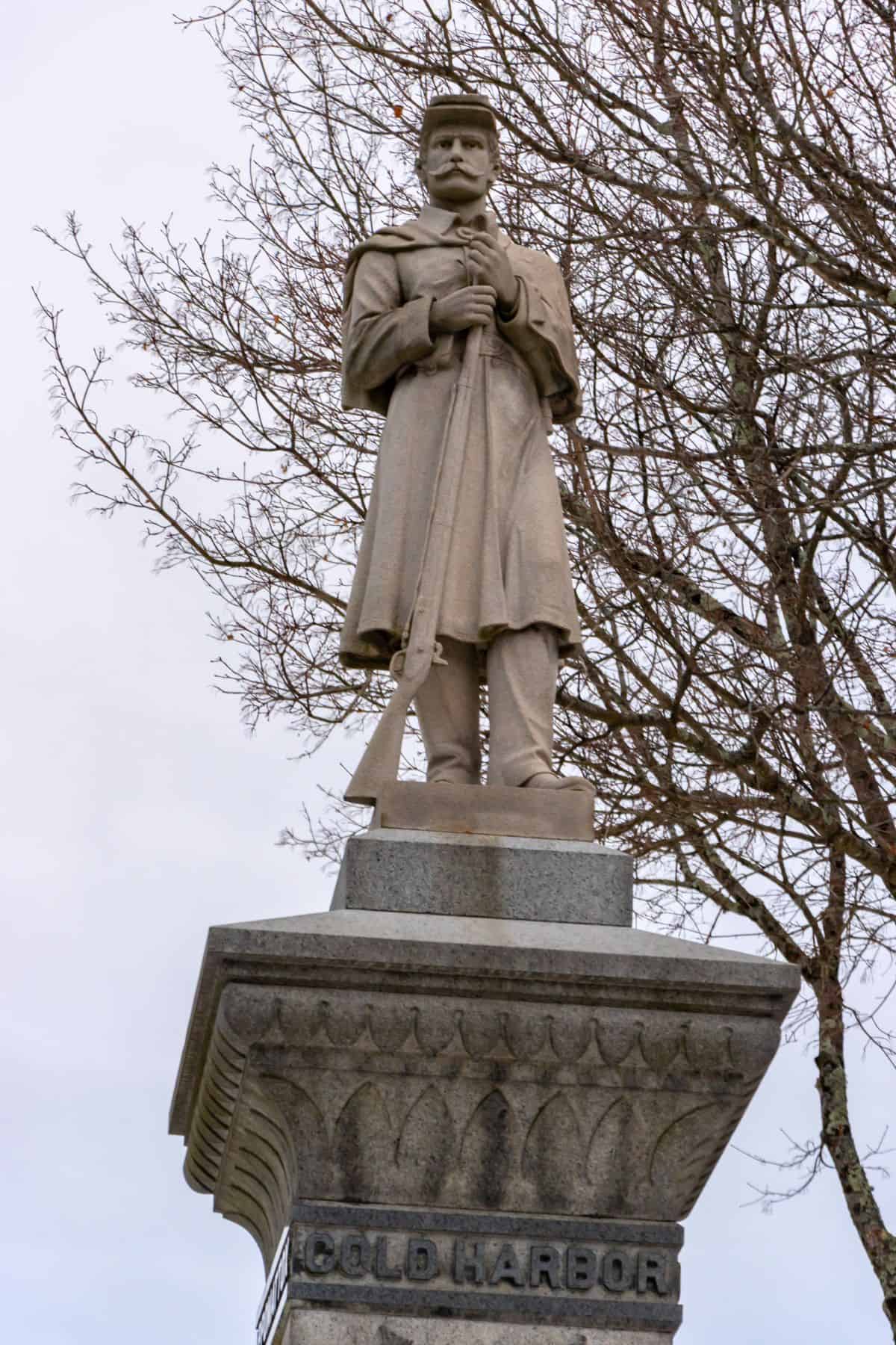 Stone statue of a soldier in uniform holding a rifle, standing atop a pedestal engraved with "COLD HARBOR," with leafless tree branches in the background.