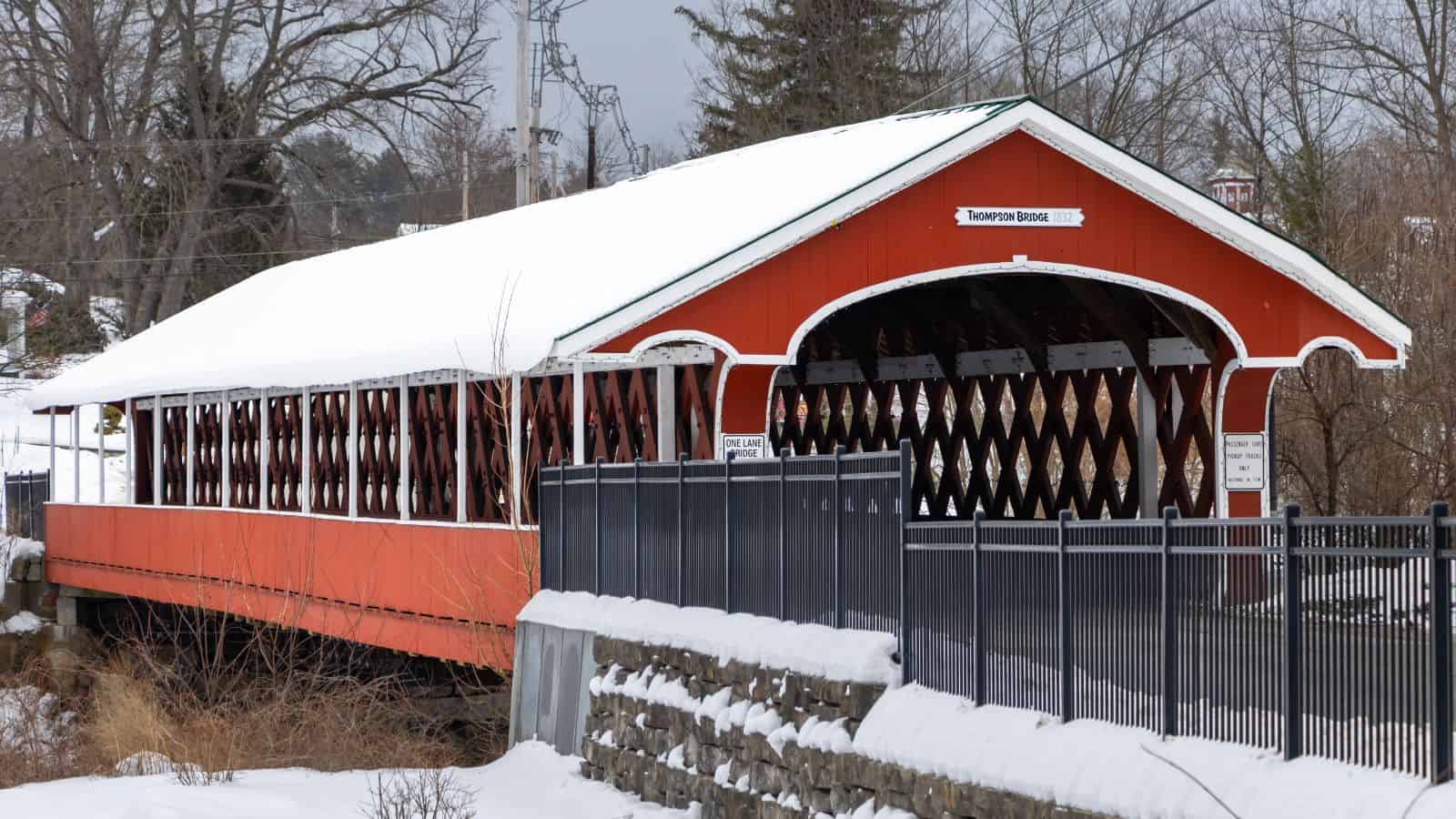 A red covered bridge with a snow-covered roof spans over a small stream, surrounded by a fence and winter scenery.