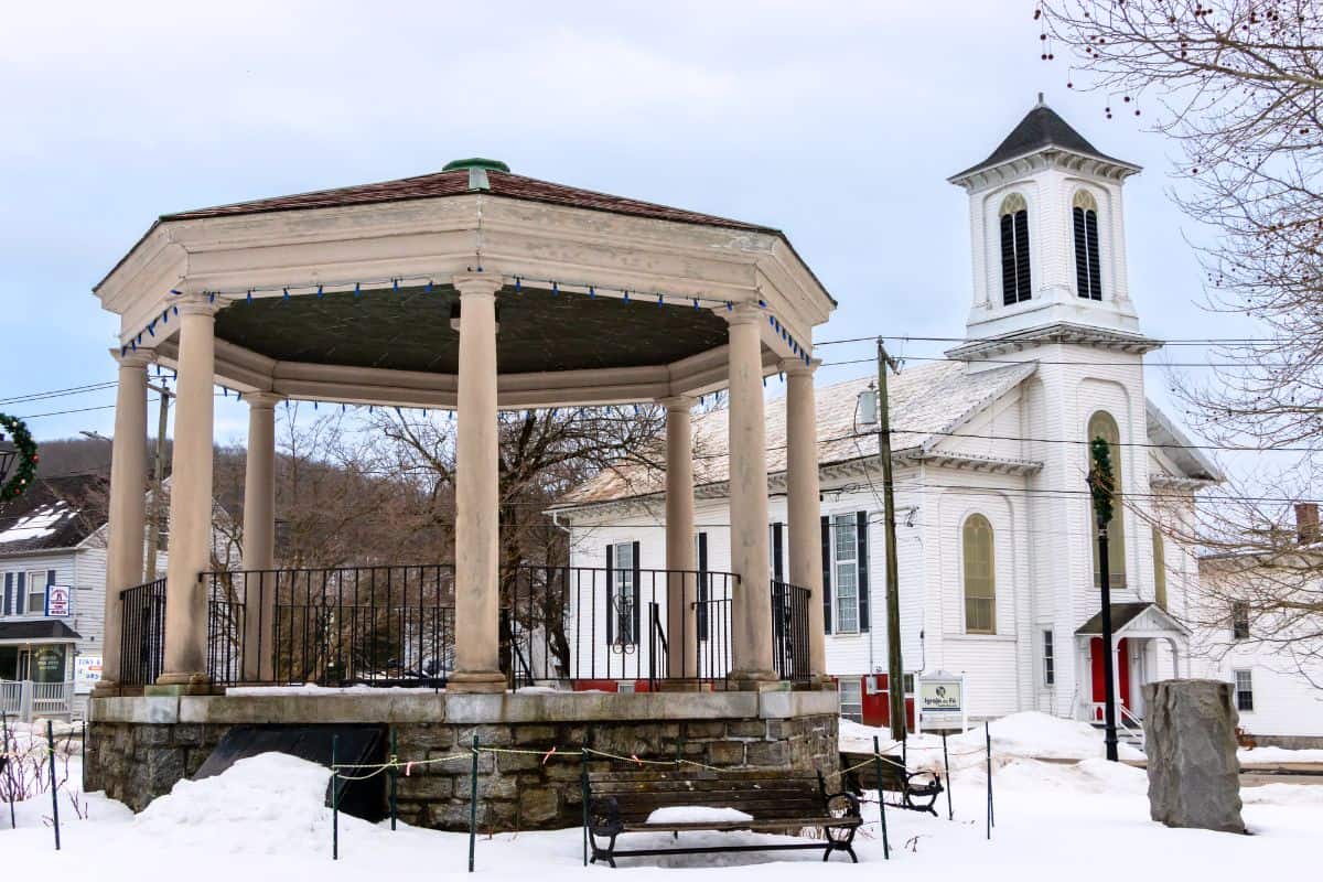 A gazebo with stone steps stands in front of a white church with a bell tower on a snowy day in a small town.