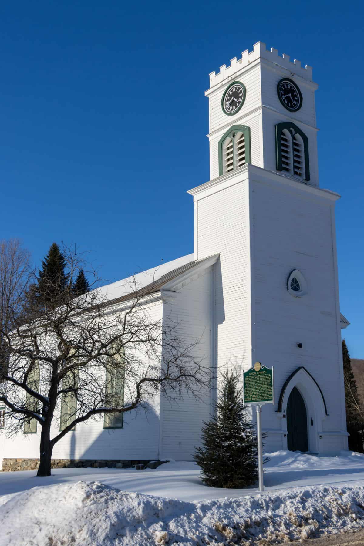 White wooden church with a tall clock tower, green-trimmed windows, and snowy ground under a clear blue sky.
