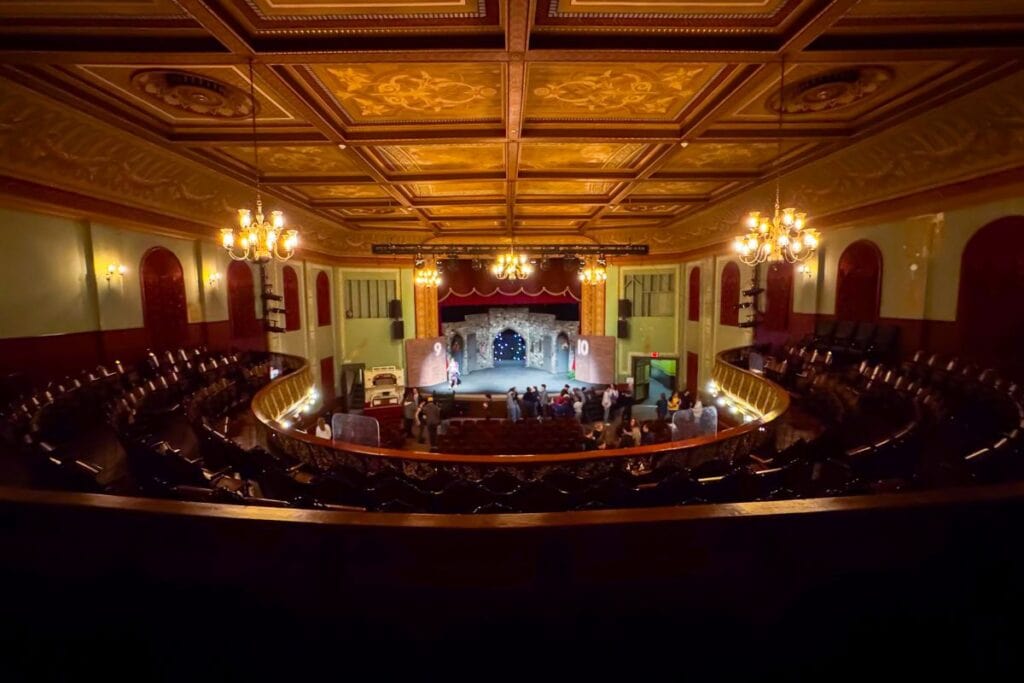 A view from the balcony of a vintage theater with ornate ceiling, chandeliers, and a stage where a performance is taking place before a small audience.