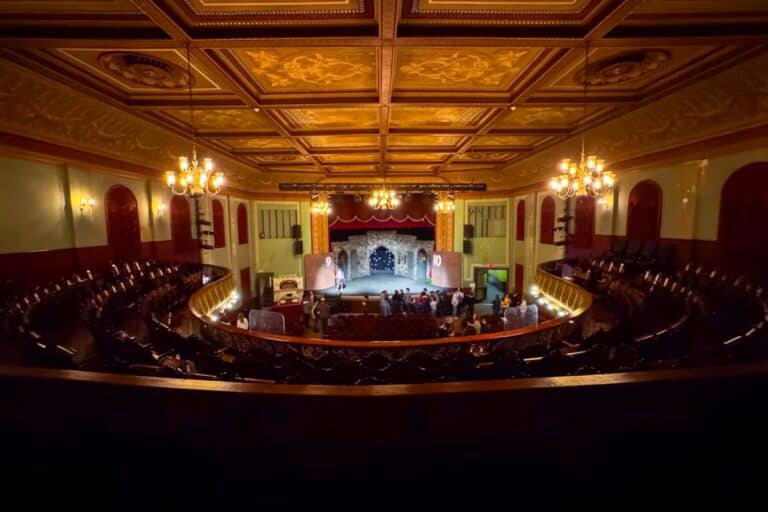 A view from the balcony of a vintage theater with ornate ceiling, chandeliers, and a stage where a performance is taking place before a small audience.