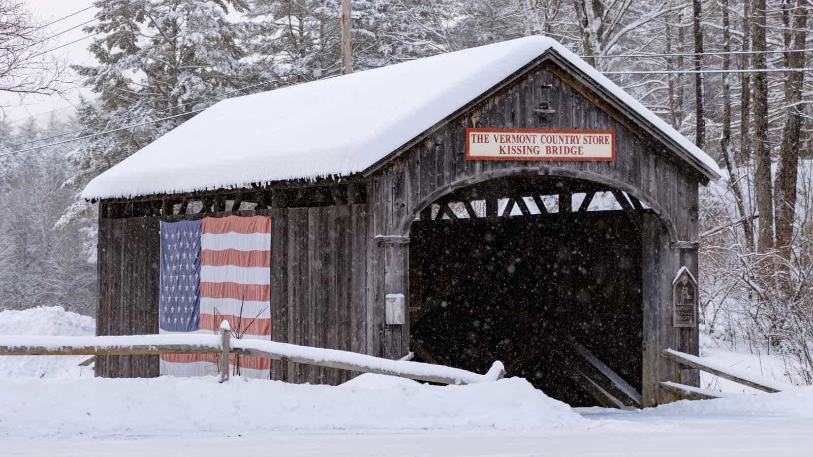 A snow-covered wooden covered bridge with an American flag hanging on the side and a sign reading "The Vermont Country Store Kissing Bridge." Snow is falling and trees are in the background.