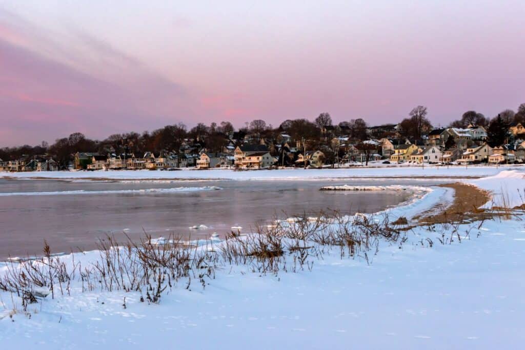 Snow-covered shoreline with frozen water in the foreground and a row of houses and leafless trees in the background under a pink and purple sky.