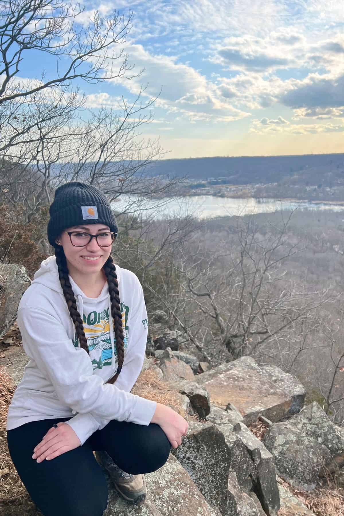 A woman with braided hair, glasses, and a beanie kneels on a rocky ledge overlooking a river and leafless trees under a partly cloudy sky.