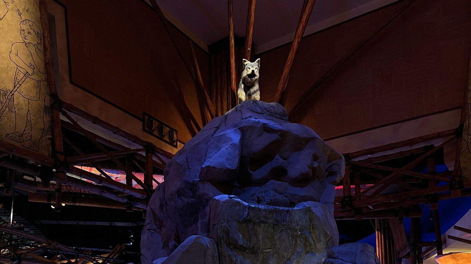 A white wolf statue stands atop a large artificial rocky formation indoors, surrounded by wooden beams and dim lighting.