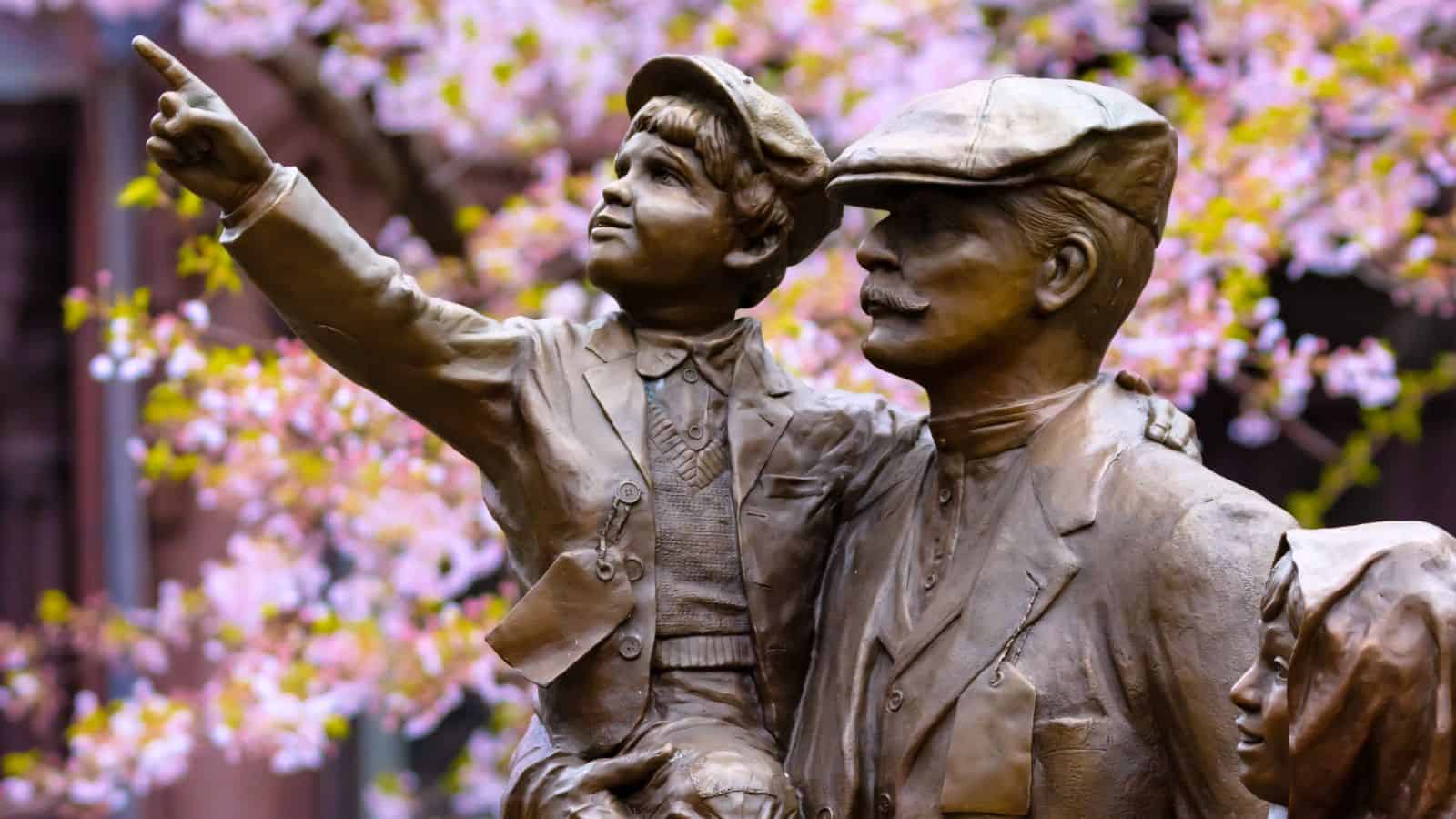 Bronze statue of a man holding a boy who is pointing, with another child partially visible; pink flowers bloom in the blurred background.