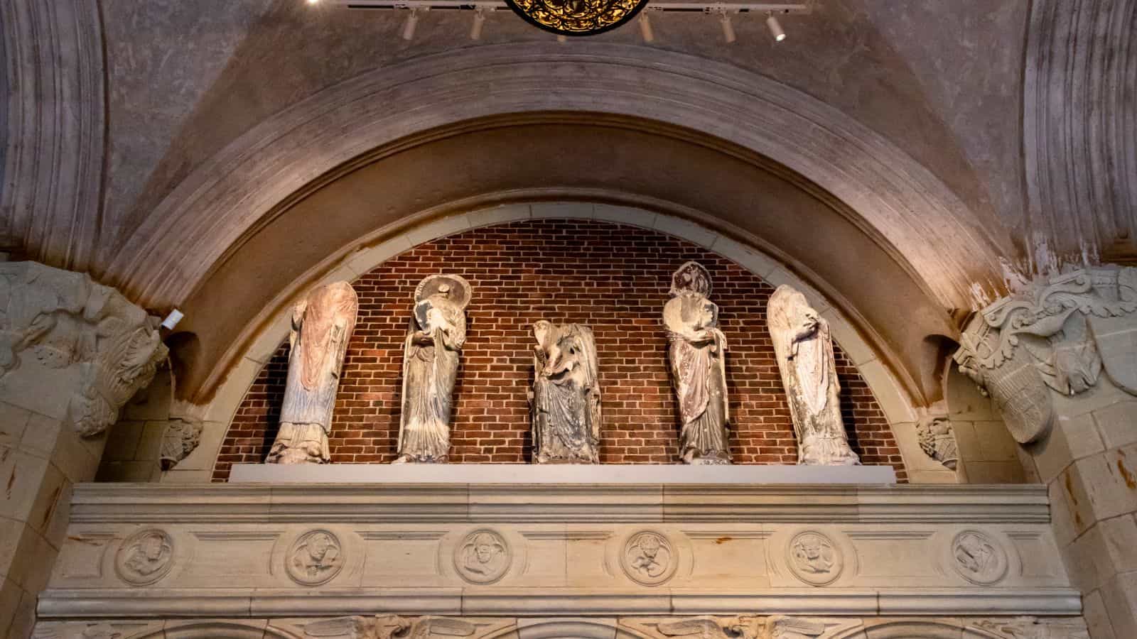 Four stone statues of robed figures are displayed in an alcove with a brick backdrop, above an ornate arch with carved medallions.
