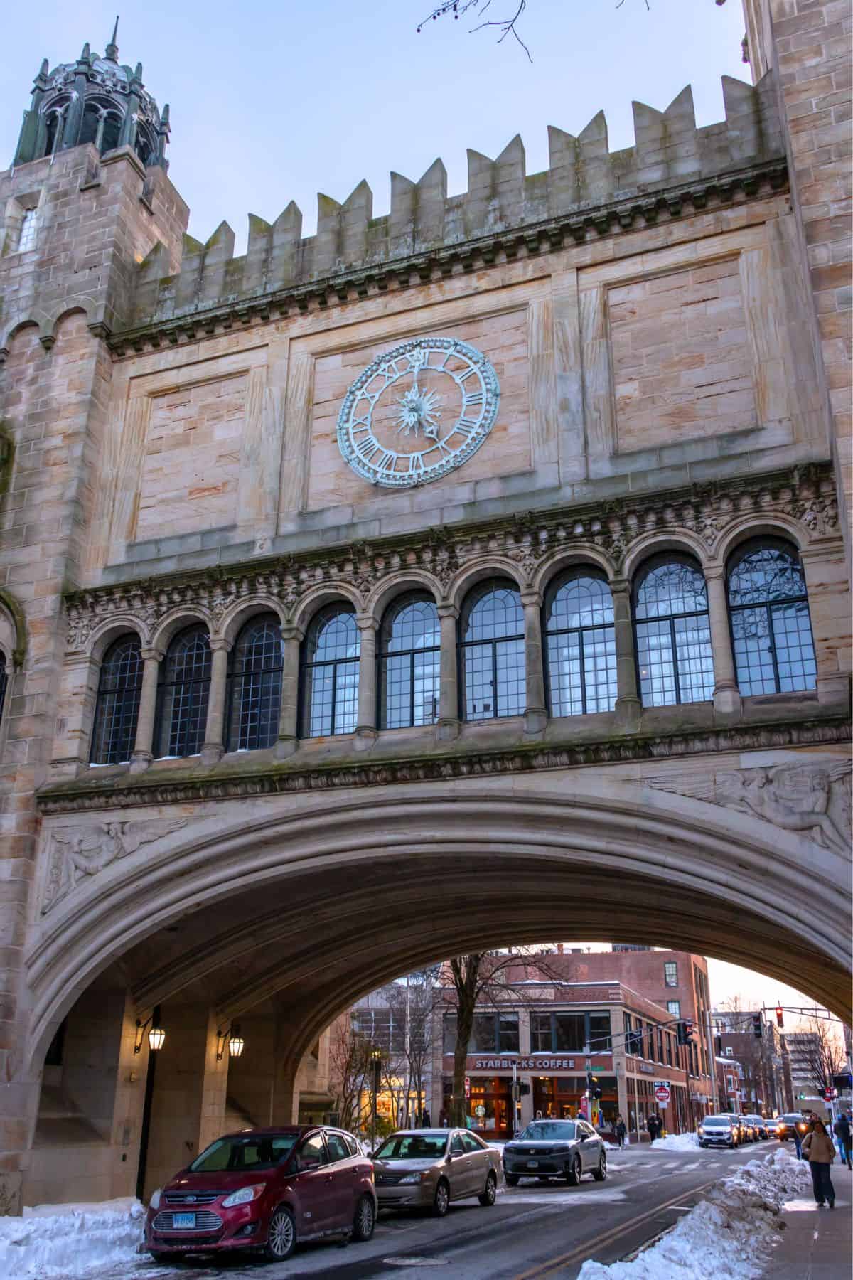 Stone archway with clock towers over a street with cars passing underneath; snow is on the ground and shops are visible in the background.