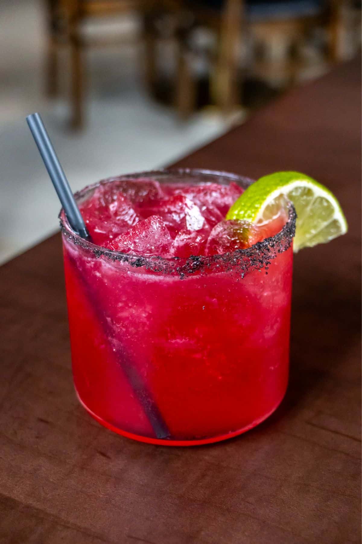 A bright red cocktail with ice, a salted rim, a lime wedge garnish, and a black straw sits on a brown table.