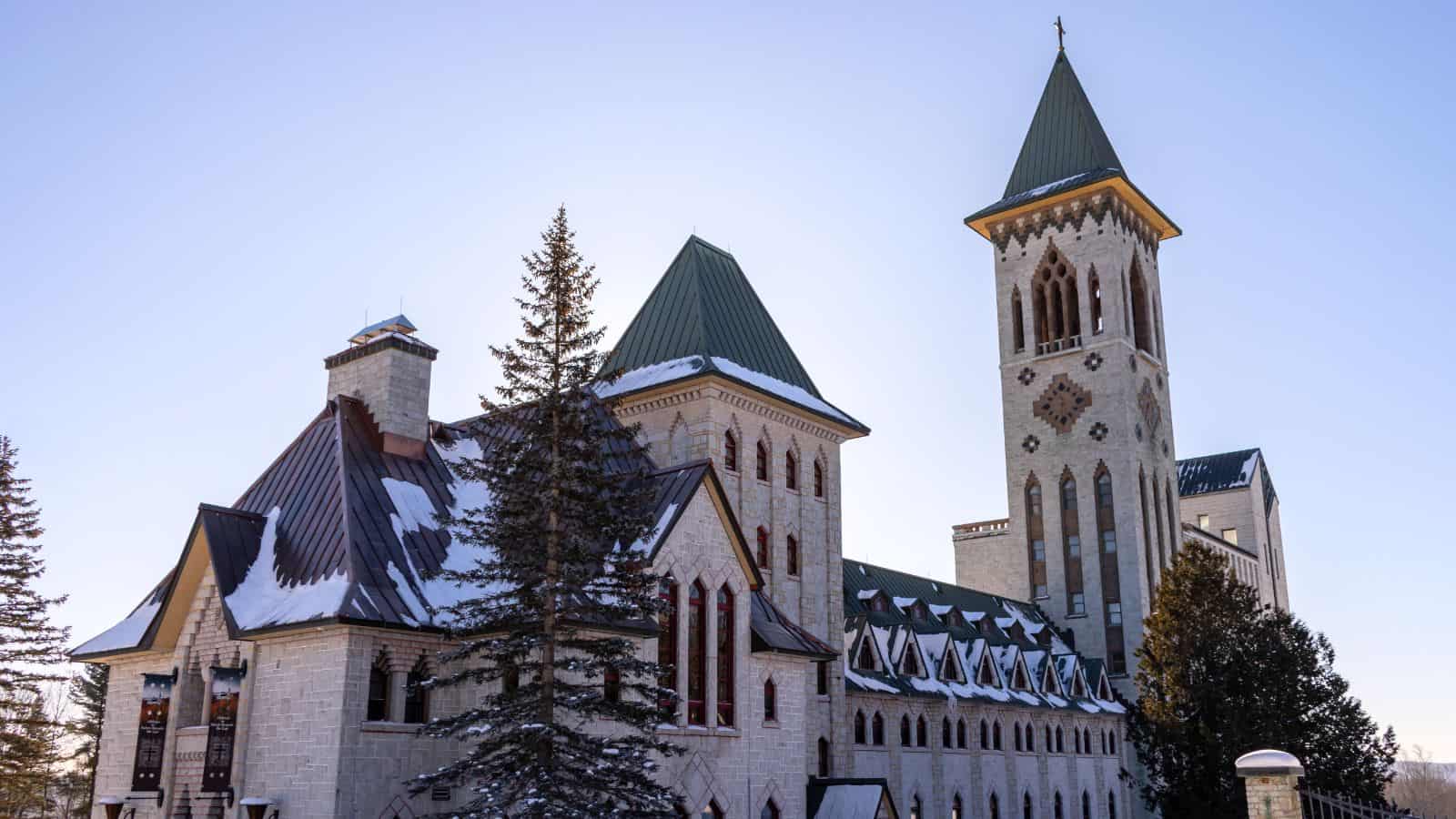 A large stone monastery with green roofs and a tall bell tower, partially covered in snow, stands among evergreen trees under a clear sky.