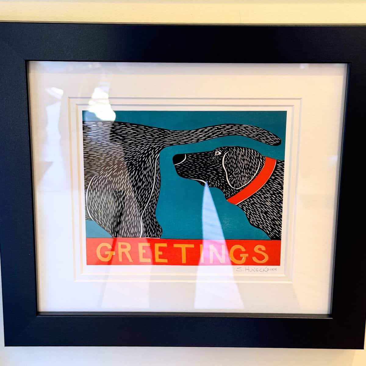 Framed print by S. Huneck featuring a black dog with a red collar on a teal background, with the word "GREETINGS" on a red banner.