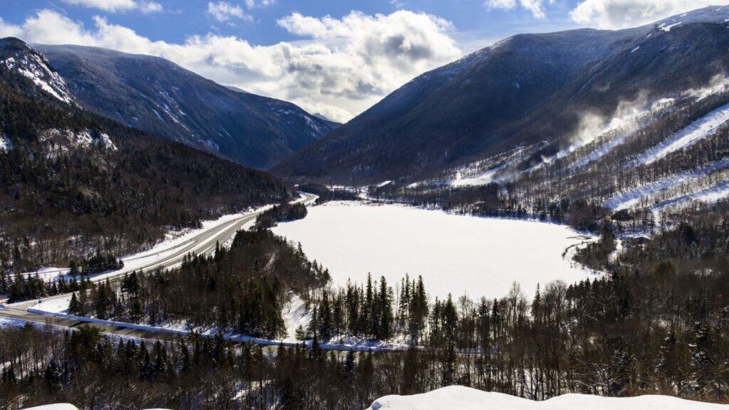 Snow-covered valley with a frozen lake, surrounded by forested mountains and winding roads under a partly cloudy sky.