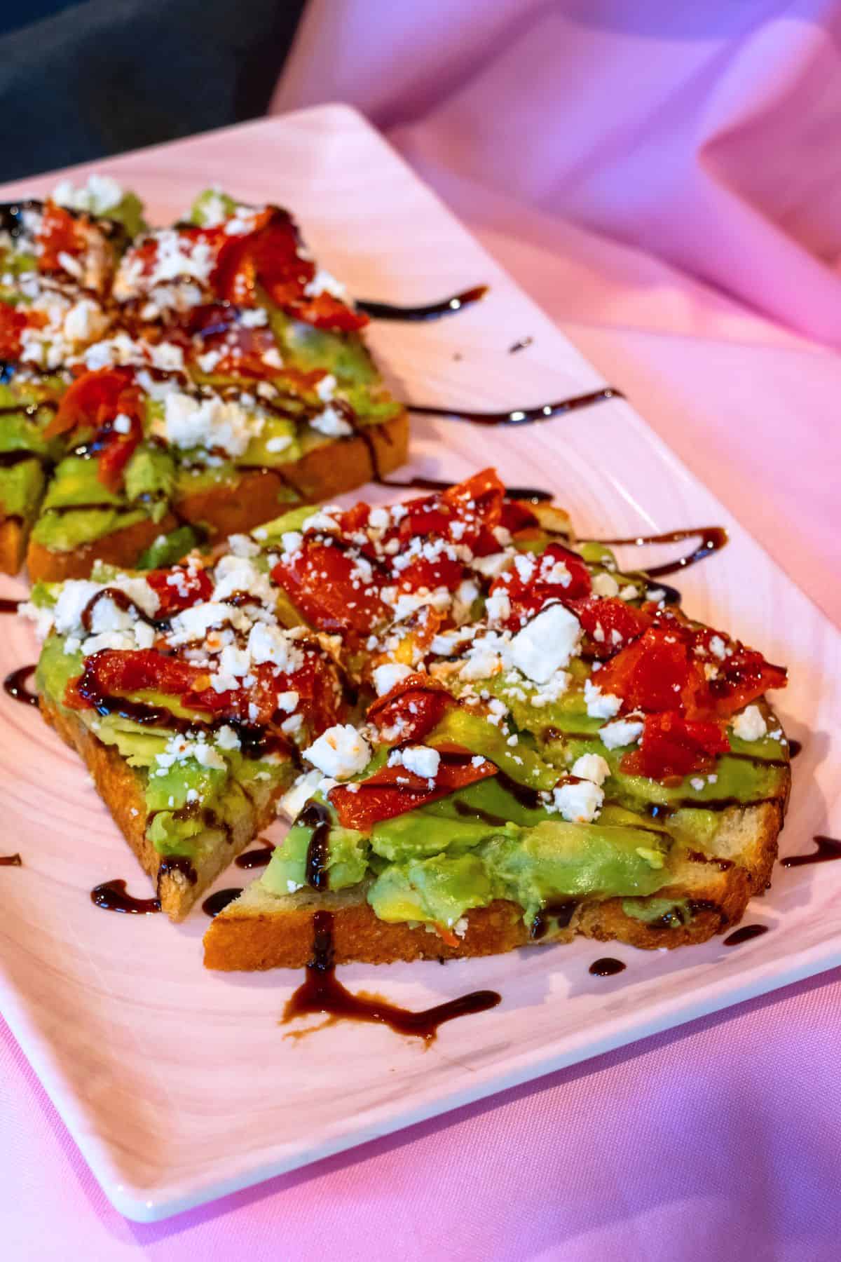 Three slices of avocado toast topped with crumbled cheese, roasted tomatoes, and a drizzle of balsamic glaze on a rectangular white plate.