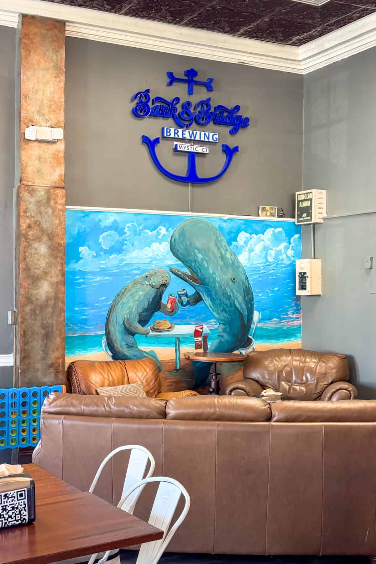 A lounge area with brown leather sofas in front of a mural of two manatees eating fast food at Bank & Bridge Brewing, with a blue neon sign above.