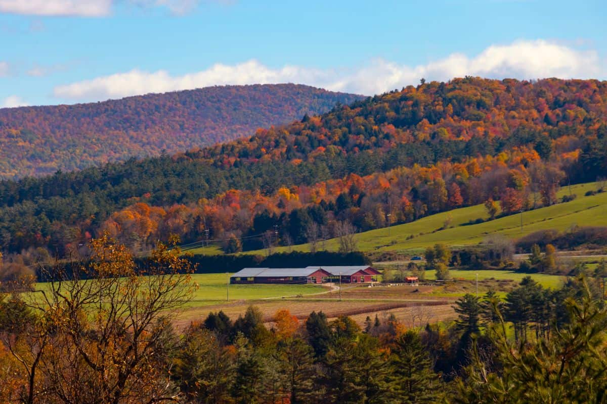 A red barn sits in a green field with rolling hills and forests of autumn-colored trees in the background under a partly cloudy sky.