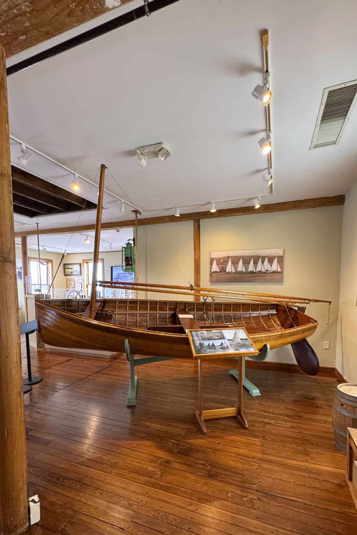 A wooden sailboat on display in a museum room, with framed boat photos on the wall and wooden floors and beams surrounding the exhibit.