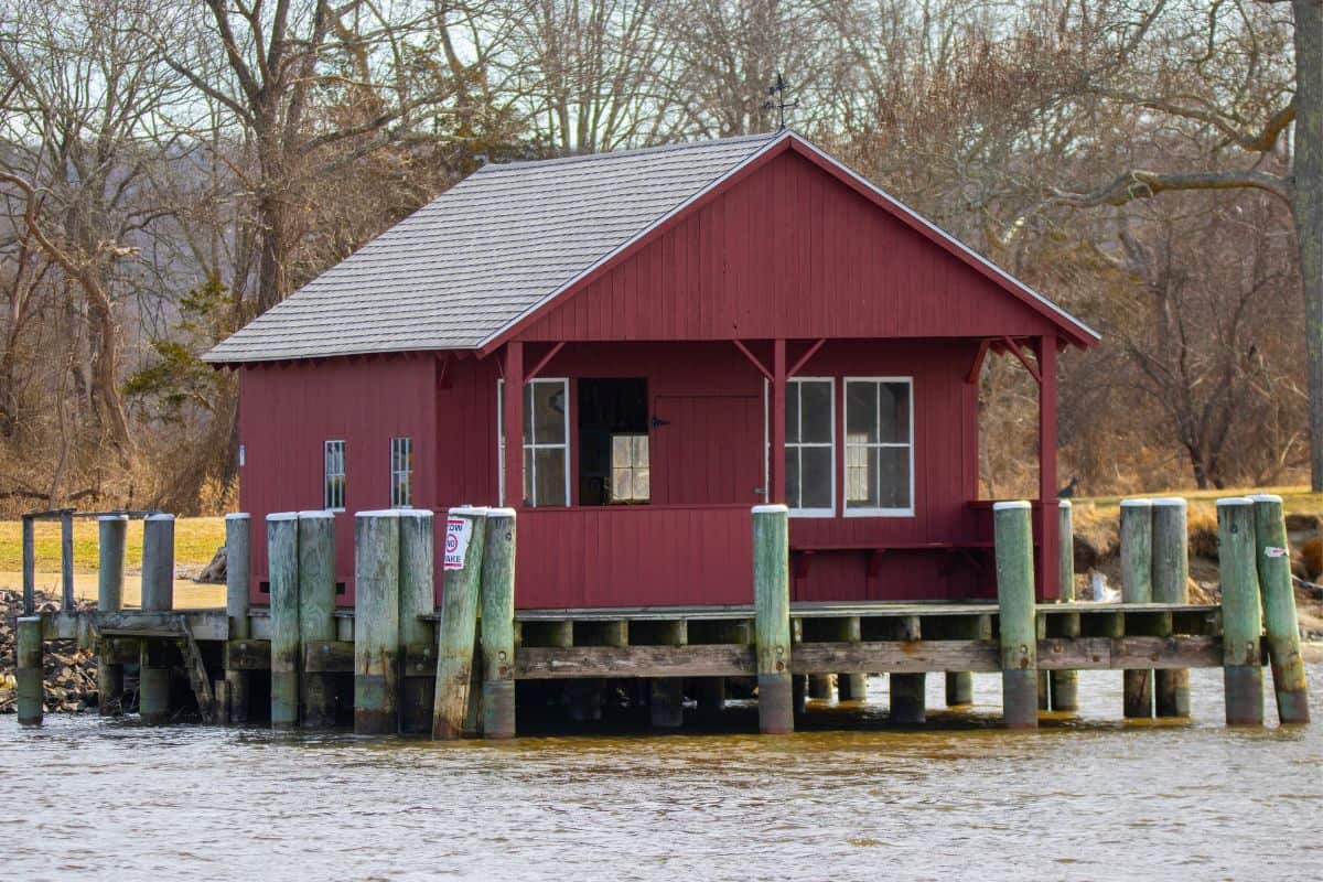 Red wooden boathouse on wooden stilts over the water, surrounded by trees with bare branches on the shore in the background.