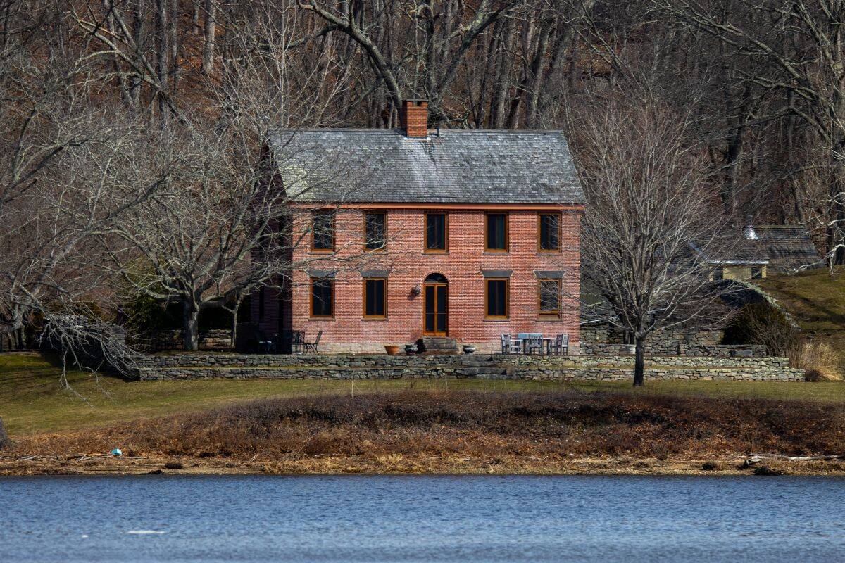 A two-story brick house with a slate roof stands behind leafless trees, facing a body of water in a rural setting.